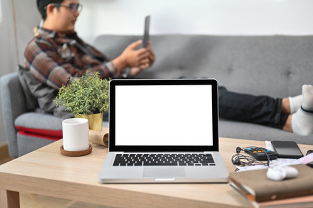 Mock up with white screen laptop putting on wooden table over a man laying on the sofa with a digital tablet as a background.の写真素材