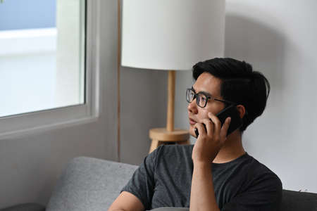 Photo of a Asain young man on a sofa is talking on a mobile phone with a comfortable living room as a background.の写真素材