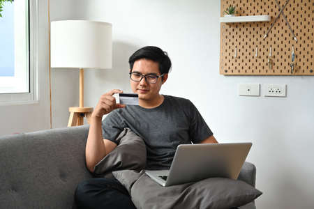 Photo of a smart man is holding a credit card while sitting with a computer laptop on the sofa.の写真素材