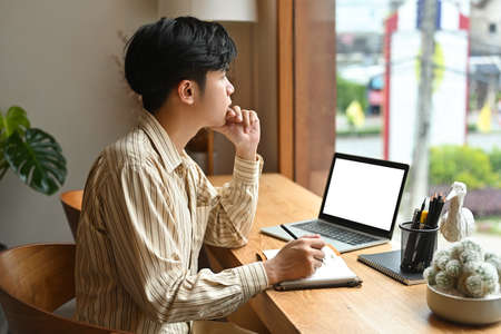 Photo of a young man keeping a hand on chin while thinking a new idea and taking notes, empty screen...