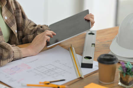 Cropped image of an architect woman's hands is holding a digital tablet at the wooden working desk.