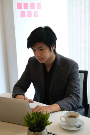 Photo of a young businessman using a laptop at the white working desk surrounded by a small potted...