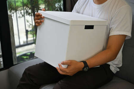 Cropped image of a young man holding a white present box while sitting on the couch in the...