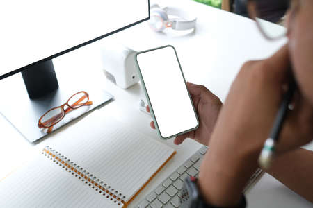 Behind view of smart man holding a white blank screen smartphone while taking notes at the working desk that surrounded by a computer monitor and various equipment.の写真素材