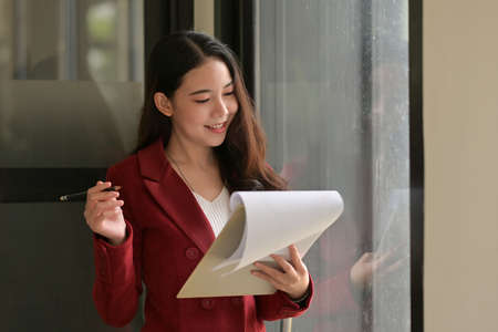 Photo of a young businesswoman standing and holding a clipboard over a comfortable meeting room as a background.の写真素材