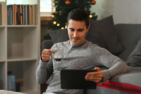 Photo of a young smart man holding a coffee cup while watching a movie from digital tablet over the comfortable couch as a background.の写真素材