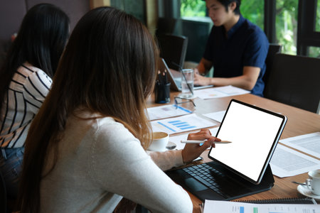Behind view image of a young designer woman using a white blank screen digital tablet and stylus pen at the long wooden table surrounded by office equipment and colleagues.の写真素材