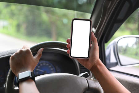 Cropped image of a hand holding a white blank screen smartphone while driving a car. Driver hand on...