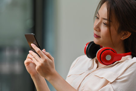 Photo of a beautiful woman with a headphones looking at the smartphone in her hand over the blur living room background.の写真素材