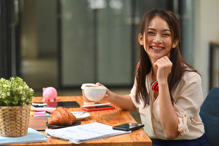 Photo of a young office woman taking a break with a coffee and croissant at the wooden working desk surrounded by paperwork, office stuff and a smartphone.の写真素材