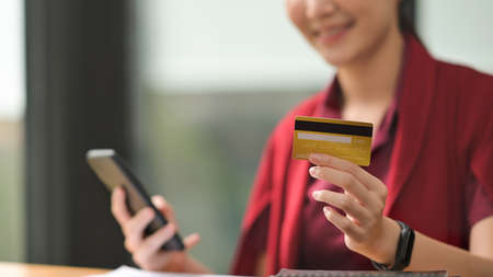 Photo of a young woman holding a credit card and smartphone in hand at the wooden working desk....