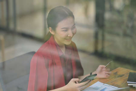 Photo through the window of a beautiful woman holding a credit card and smartphone at the wooden working desk.の写真素材