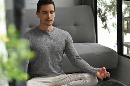 Photo of a young man with wireless earphones doing yoga and meditating on the living room floor over...