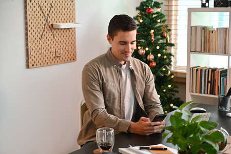 Photo of a young man using a smartphone at the working desk over the Christmas tree and bookshelf in...