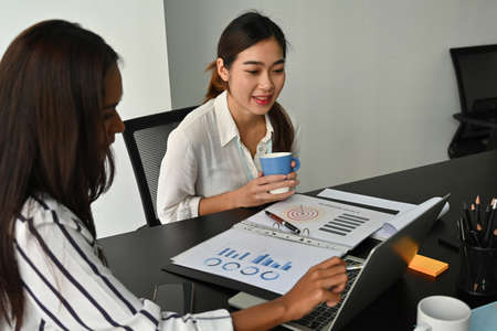 Photo of businesswoman drinking coffee while sitting with a colleague at the working desk surrounded by office equipment.の写真素材