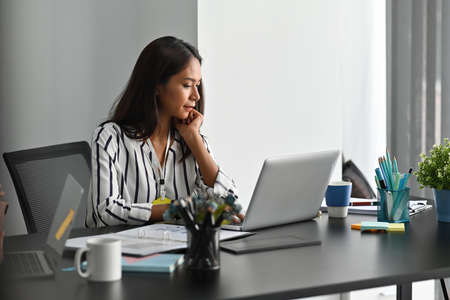 Photo of a young business owner looking at the computer laptop at her modern working desk surrounded...