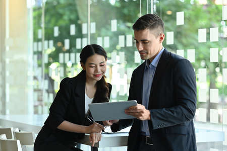 Photo of a young businessman using a digital tablet and stylus pen while standing together with the secretary over the modern office as a background.の写真素材