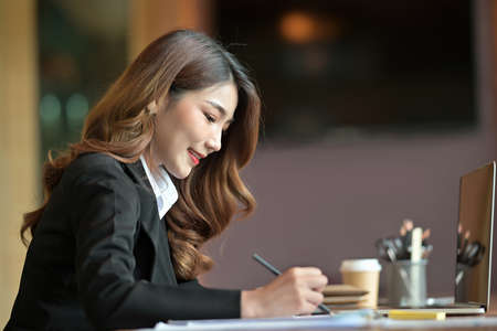 Photo of a beautiful businesswoman taking notes at the working table surrounded by a computer laptop and office equipment.の写真素材