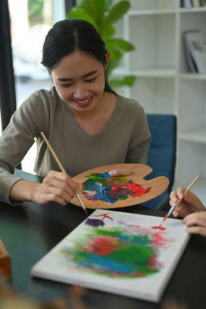 Photo of a young teacher holding a palette and drawing together with her student at the student desk.の写真素材