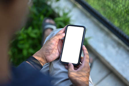 Top view image of a man's hand holding a white blank screen smartphone while sitting next to the wire mesh fence.の写真素材