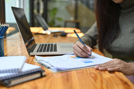Cropped image of a young woman writing on paperwork while sitting at the wooden working desk surrounded by a computer laptop and office equipment.の写真素材
