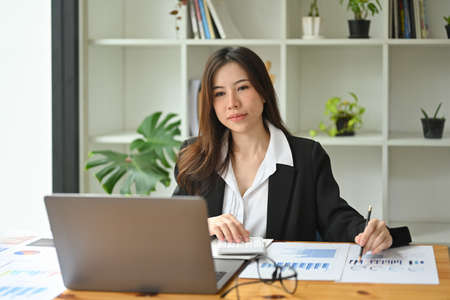 Photo of a beautiful insurance agent using a computer laptop at the wooden working desk surrounded...