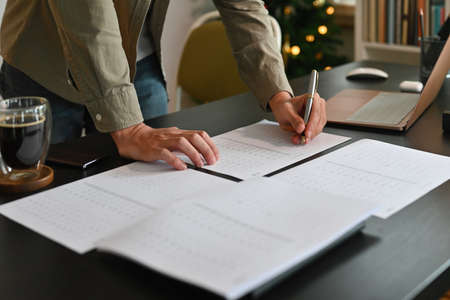 Close-up image of a man standing and leaning on a working desk surrounded by paperwork, computer...