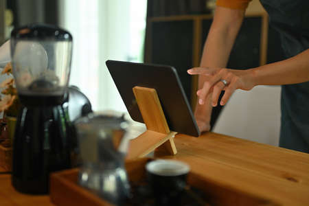 Photo of barista's hand using a digital tablet at the wooden counter surrounded by coffee equipment.