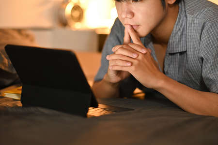 Photo of a young man in pajamas lying on the bed and using a digital tablet.