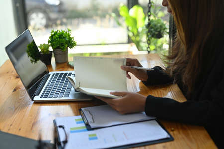 Cropped image of an office woman taking notes at the wooden table surrounded by a computer laptop,...