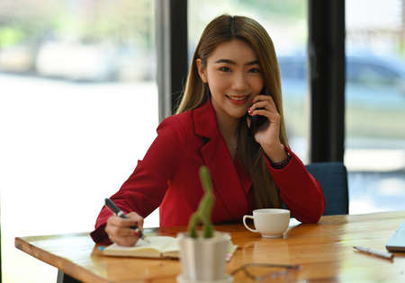 Photo of a businesswoman talking on mobile phone at the wooden table surrounded by a computer laptop and various office equipment.の写真素材