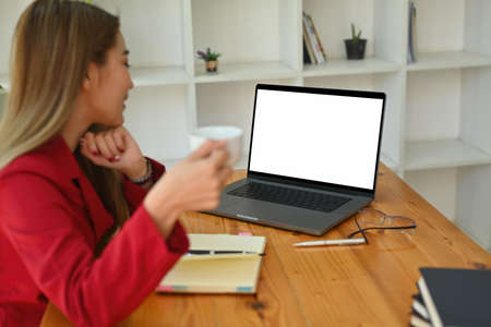 Photo of a white blank screen computer laptop on the wooden table surrounded with blur office woman...