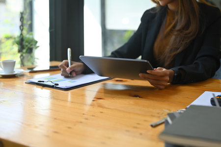 Cropped image of a businesswoman writing on a clipboard and holding a digital tablet at the wooden working desk.の写真素材