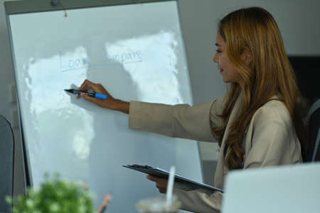 Photo of female banker writing on the board while standing and holding a clipboard in the comfortable meeting room.の写真素材