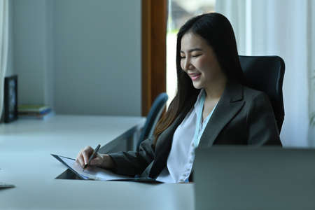 Photo of a young businesswoman writing on a clipboard while sitting at the modern working desk.の写真素材