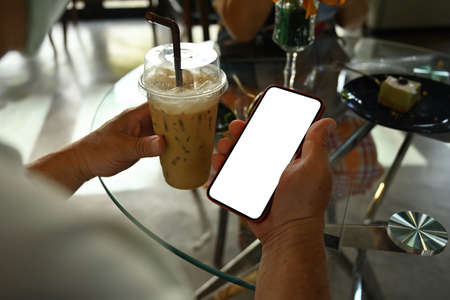 Photo of a man with medical mask holding an iced coffee and using a white blank screen smartphone at the glass table.の写真素材