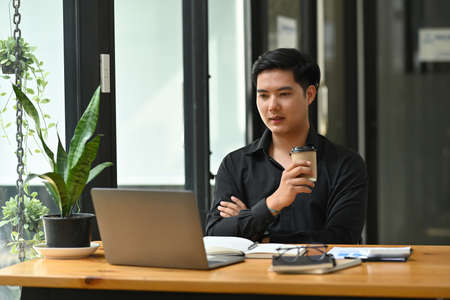 Photo of a young smart office man holding a takeaway coffee cup and looking at the computer laptop at the wooden working desk.の写真素材