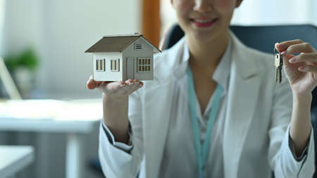Photo of a young real estate broker holding a house model and showing a key at the modern working desk.の写真素材