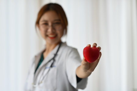 A red heart model holded by Asian woman physician with glasses as over blur background.