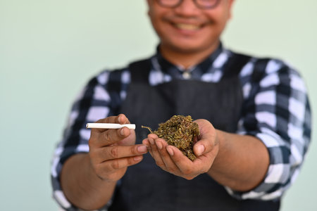 Asian man presenting marijuana tobacco and other hand holding dried marijuana flower, feeling high...