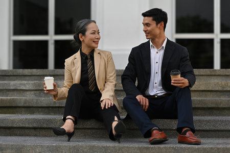 Coffee break and chill out after work with Senior businesswoman and Young smart businessman sitting on the stairs in front of office.の写真素材