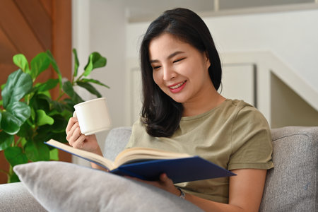 Millennial Asian girl sitting on sofa and enjoy with morning coffee and  book at home.の写真素材