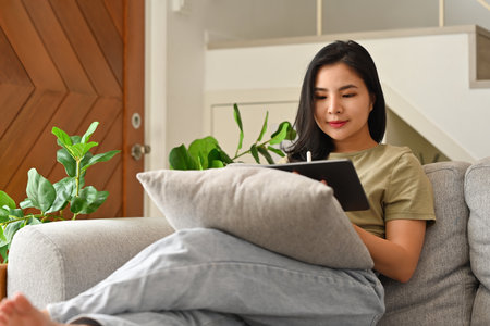 Calm Young Asian woman sitting on the sofa and reading a book at home.の写真素材