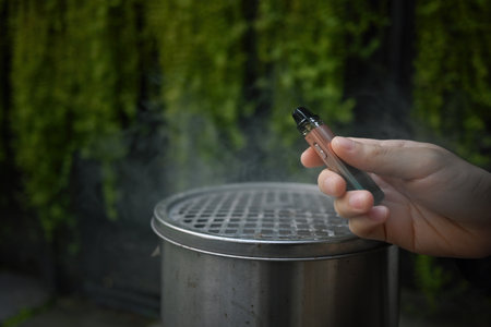Close up with male's hand holding e-cigarette and empty ashtray on the background.