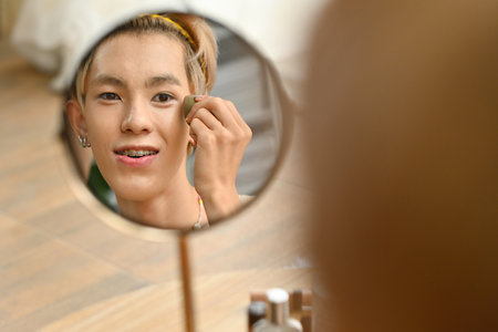 Photograph reflected in the mirror with Young Asian gay teenage using a makeup sponge to blend...