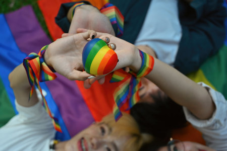 Above view with group of LGBT teenagers lying down on rainbow flag and holding heart model together,...