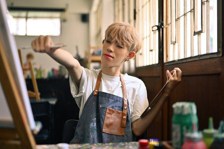 Portrait of a young LGBT artist smiling with colored hair holding a paintbrush and pointing to the...