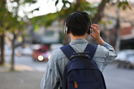 Rear view of Asian backpacker male walking on footpath while listening a music from headphones in the urban city, Solo travel concept.の写真素材