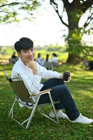 Vertical image of a Young Asian man relaxing on a camping chair with coffee drink.の写真素材