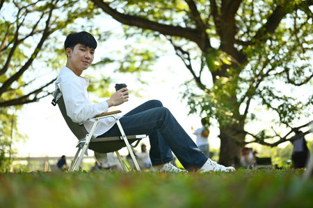 Side view image of a Young Asian man relaxing on a camping chair with a coffee drink.
